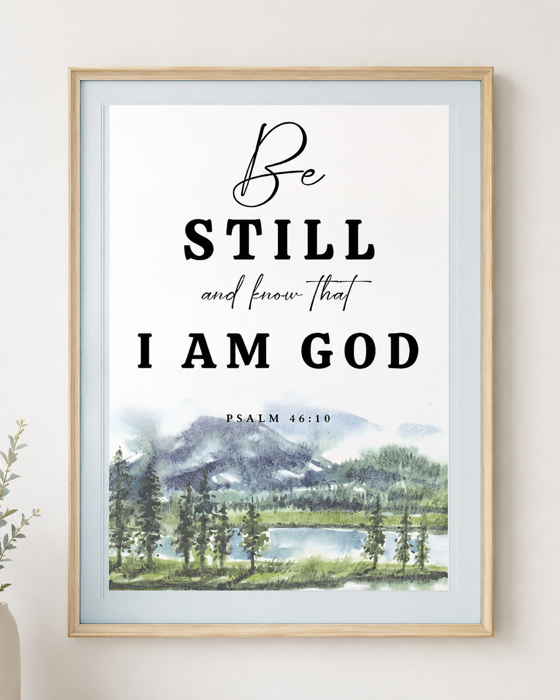 Framed Psalm 46:10 scripture print with bold mixed typography and a watercolor mountain lake landscape, hung above a light oak bed with blue and cream bedding in a calm bedroom.
