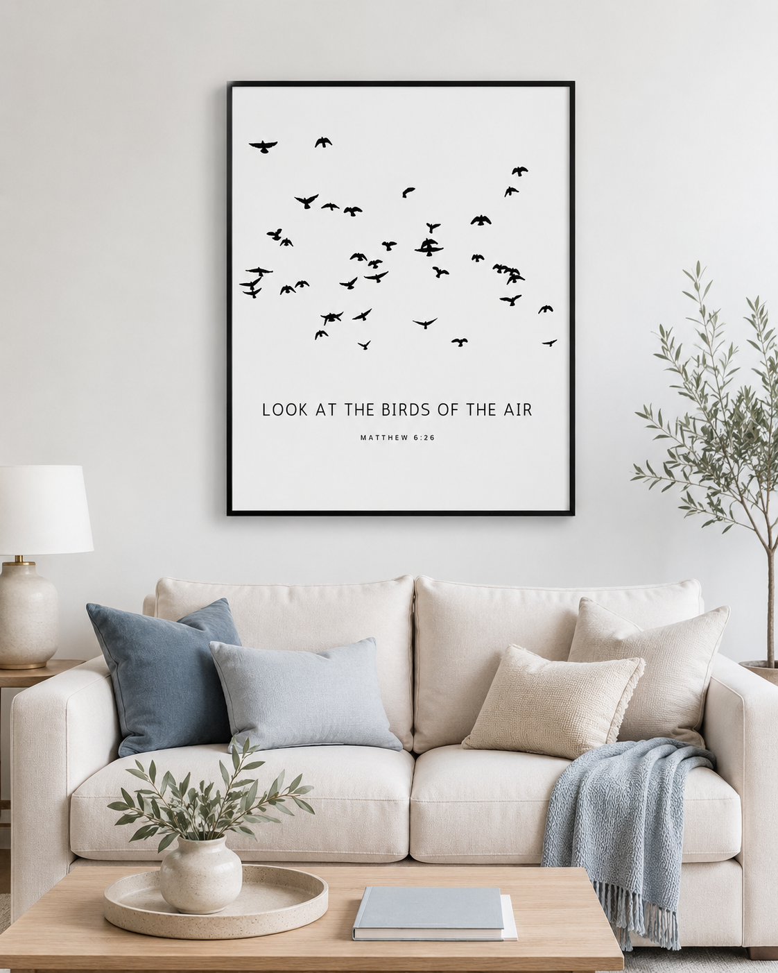 Large square black-framed wall art print of a flock of birds with Matthew 6:26 scripture text, hung above a cream linen sofa in a bright minimalist living room.
