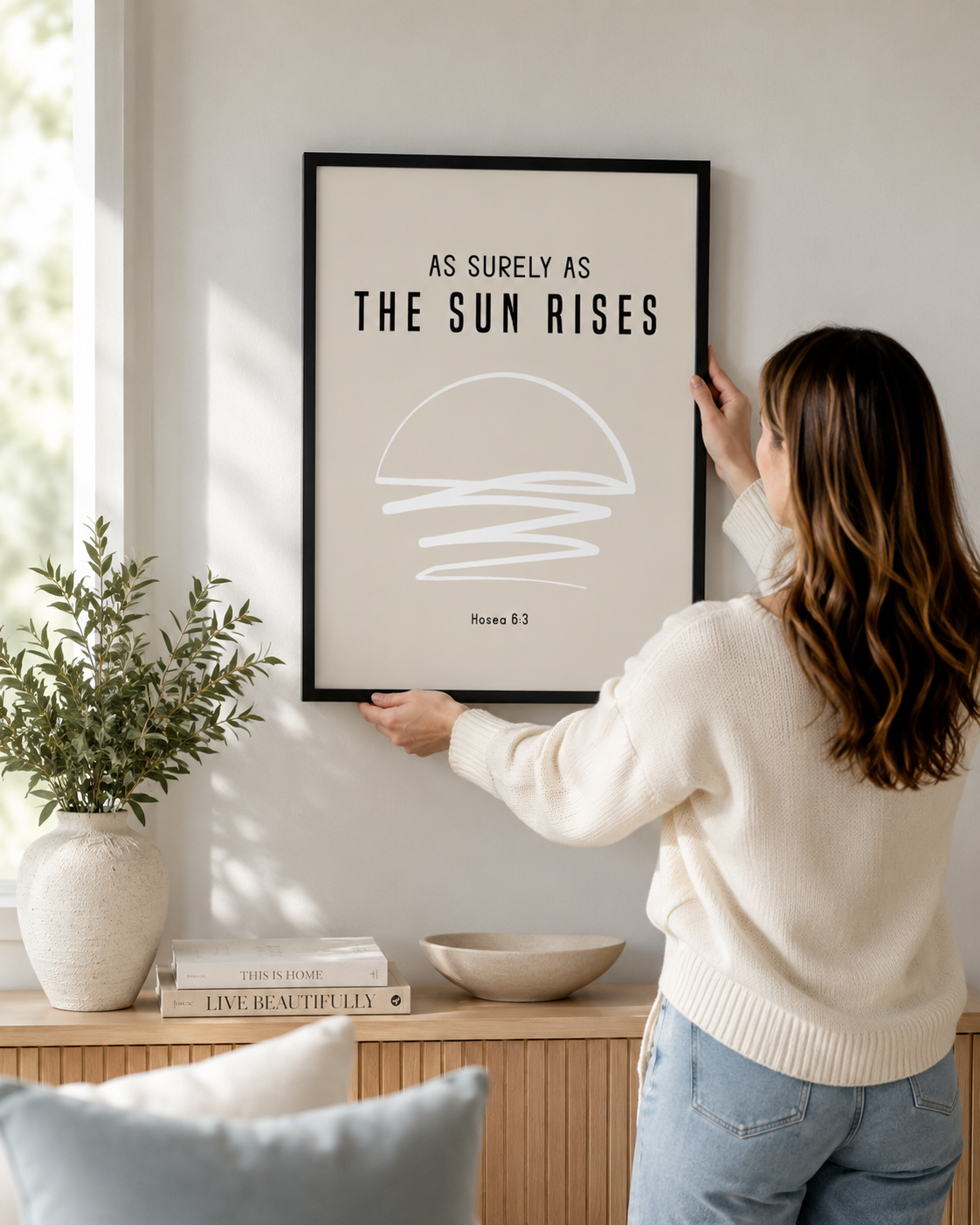Woman hanging a framed Hosea 6:3 "As Surely As the Sun Rises" print on a wall
