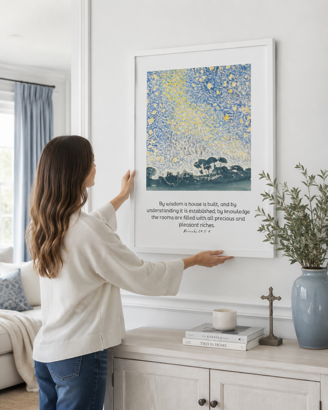 Woman hanging a framed Proverbs 24:3-4 "By Wisdom a House Is Built" print on a wall