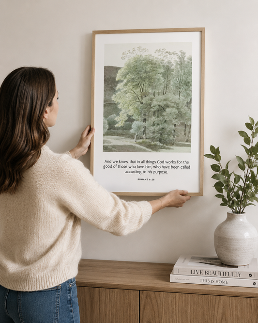 Woman hanging a framed Romans 8:28 Bible verse print above a sideboard at home