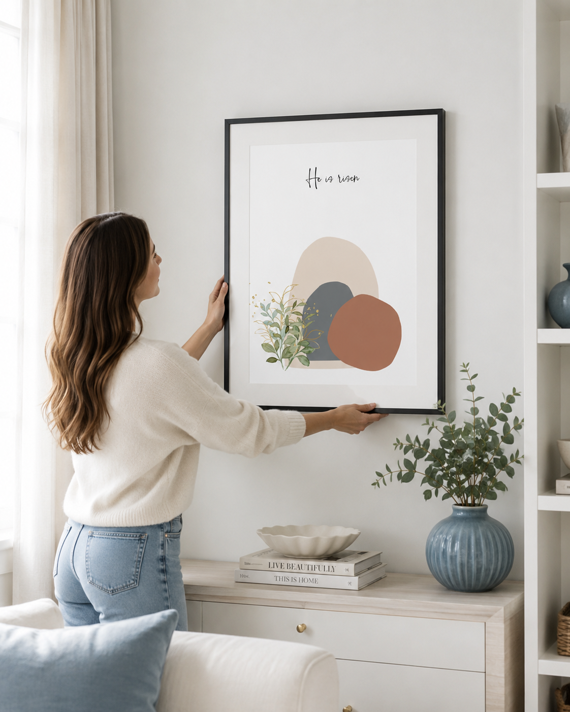 Woman hanging a He Is Risen Easter wall art print on a living room wall